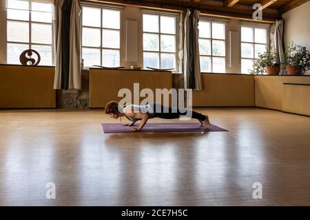 Ganzkörper-Seitenansicht der konzentrierten Frau in Sportbekleidung üben Shakti Yoga allein und stehend in Plank Position in geräumig studio Stockfoto