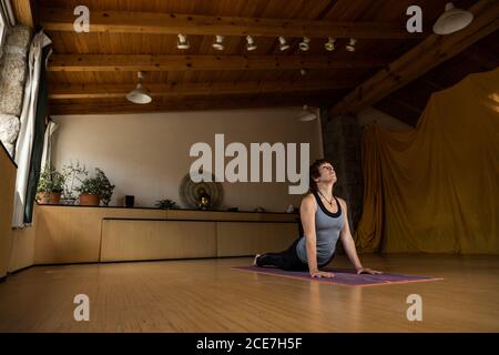 Ganzkörper-Seitenansicht der konzentrierten Frau in Sportbekleidung üben Shakti Yoga allein dabei Kobra Pose in geräumigen Studio Stockfoto