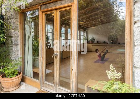 Seitenansicht einer nicht erkennbaren Frau in Sportkleidung, die im Boot sitzt Pose beim Üben Shakti Yoga allein im Lichtstudio mit Holzinterieur in tropischem Resort Stockfoto