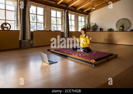 Ruhige Frau in Sportbekleidung sitzt auf Matte in geräumigen Studio Und Yoga in Padmasana üben, während ein Online-Tutorial gedreht wird Auf einem Laptop Stockfoto
