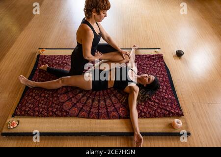 Von oben: Weibliche Trainerin, die der flexiblen Frau beim Strecken der Beine in Eka Pada Pavanamuktasana hilft, während sie im Studio Yoga macht Stockfoto