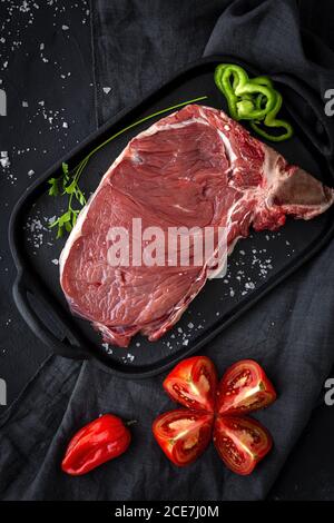 Frisches rohes T-Bone-Rindersteak mit rotem Pfeffer, Tomaten, rosa Pfeffer und Petersilie, fertig zum Kochen von oben. Flach liegend. Draufsicht Stockfoto