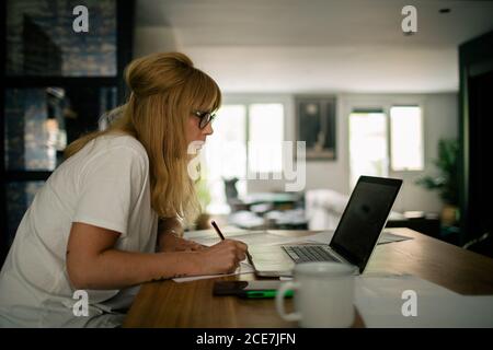 Seitenansicht der ernsthaften weiblichen Unternehmer in lässiger Kleidung sitzen Am Tisch mit Netbook in der modernen Wohnung und arbeiten an Projekt während des Schreibens auf Papier Stockfoto