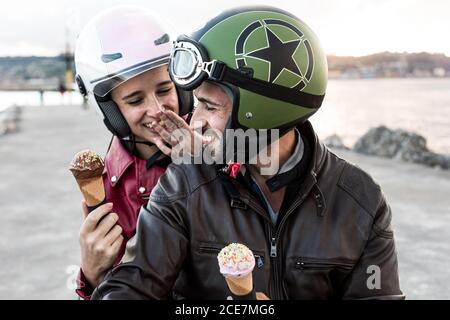 Glücklicher bärtiger Mann in Helm und Lederjacke mit Freundin Essen lecker Eis Kegel, während auf dem Fahrrad in der Nähe sitzen Fluss unter bewölktem Himmel Stockfoto