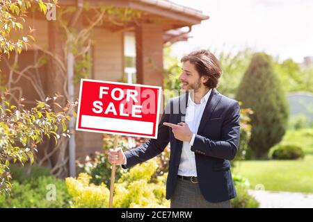 Zuversichtlich Immobilienmakler zeigt auf Zeichen mit Worten FÜR VERKAUF vor dem Wohnhaus draußen Stockfoto