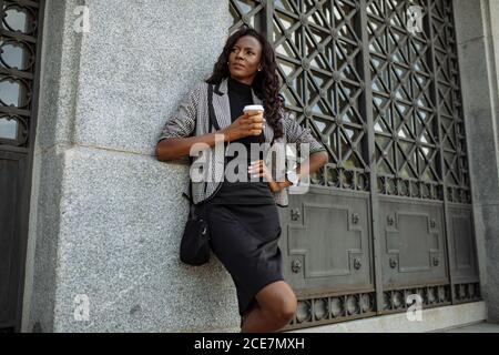 Ein niedriger Blickwinkel einer selbstbewussten jungen, gut gekleideten ethnischen Geschäftsfrau mit einer Tasse Kaffee zum Mitnehmen, die sich an der Steinmauer des Stadtgebäudes zurücklehnt und während des Arbeitstages eine Pause macht Stockfoto