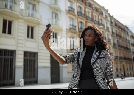 Selbstbewusste junge afroamerikanische Geschäftsfrau in stilvollem formellen Outfit, die auf der Stadtstraße steht und Selfie auf dem Smartphone nimmt Stockfoto