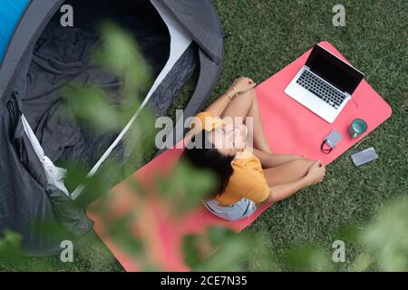 Draufsicht auf entspannte junge Frau im lässigen Outfit sitzend In der Nähe Zelt mit Laptop und Smartphone und genießen Sie das Sommerwochenende Auf grüner Wiese Stockfoto
