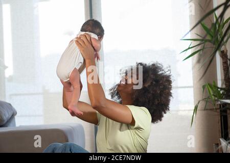 Seitenansicht einer entzückten afroamerikanischen Frau, die ein niedliches Kleinkind wirft, während sie das Wochenende zu Hause genießt Stockfoto