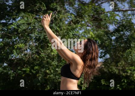 Seitenansicht der glücklichen jungen Frau im Bikini unter stehen In der Nähe von grünen Bäumen Selfie auf dem Smartphone zu nehmen, während Spaß Am Pool im Sommer Tag Stockfoto