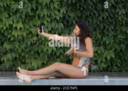 Seitenansicht der positiven jungen Frau in stilvollen Bikini und Sonnenbrillen sitzen mit niedlichen Hund am Pool gegen grüne Pflanzen Und genießen Sie den Sommertag, während Sie ein Selfie mit dem Smartphone machen Gemeinsam Stockfoto