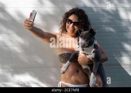 Fröhliche junge Frau in Bikini und Sonnenbrille hält niedlichen Hund Und Selfie mit dem Handy machen, während man gegen Weiß steht Wand während des Sommertages am Pool Stockfoto