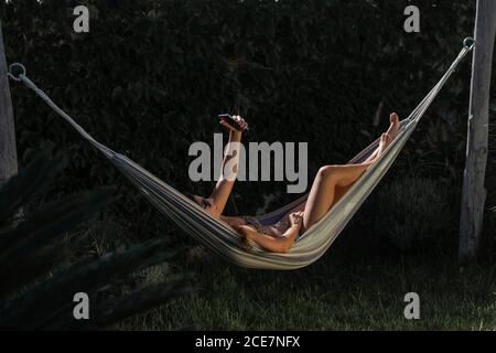Seitenansicht der fröhlichen jungen Frau in Bikini und Sonnenbrille Liegen in der Hängematte in der Nähe von grünen Pflanzen und Selfie auf Smartphone während der Erholung im Sommer Tag im Garten Stockfoto