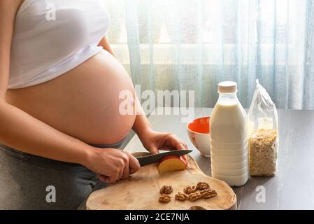 Junge und schöne schwangere Frau bereitet gesundes Frühstück zu Hause. Zugeschnittenes Bild von schwangeren Frau Kochen ein Frühstück mit Haferflocken, Apfel, mi Stockfoto