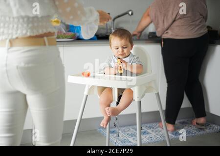 Fokussiertes kleines Kleinkind sitzt auf Baby-Hochstuhl und untersuchen Frische Avocado, während Mutter Essen in der heimischen Küche zubereitet Stockfoto