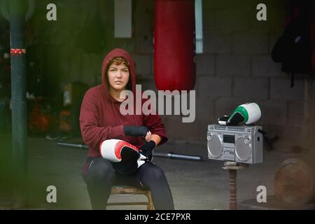 Konzentrierte Boxerin in Leggings und Kapuzenjacke, die Hände einwickelt Black Boxing Wraps vor dem Boxen Training in Shabby Keller Fitness-Studio Stockfoto