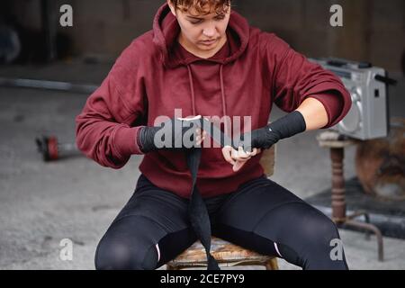 Crop konzentrierte weibliche Boxerin in Leggings und Kapuzenjacke Wickelhands Mit schwarzen Boxpackungen vor dem Boxtraining im schäbigen Keller Fitnessraum Stockfoto