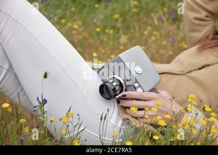 Seitenansicht von schlanken anonymen weiblichen Fotografen in Jeans Abdeckung Gesicht mit Hut beim Liegen auf Wiese mit bunten Blumen Und halten alte Videokamera in Land Stockfoto