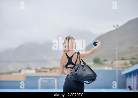 Rückansicht der unkenntlich schlanken Frau in Sportbekleidung mit Sport Tasche über der Schulter stehen auf Sportplatz und Selfie Auf dem Handy nach dem Training im Sommer Tag Stockfoto