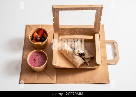 Von oben köstliche Buchweizenkrepe mit gebratenem Gemüse in Kartonbehälter mit Rote Bete Sahne und reifen Früchten in angeordnet Schüsseln auf dem Tisch Stockfoto