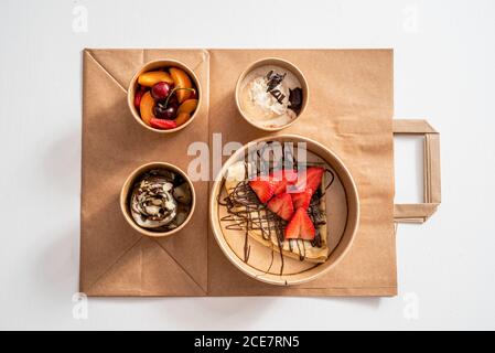 Blick von oben auf verschiedene Eiscreme mit Schokolade und dekoriert Mandelblüten in der Nähe verschiedene frische Früchte und Buchweizen Crepe mit Erdbeerscheiben in umweltfreundlichen Schachteln auf dem Takeaway-Beutel Stockfoto