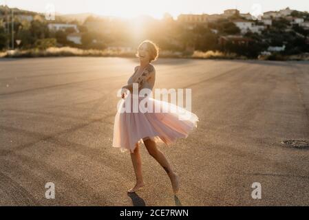 Ganzkörper begeistert junge Künstlerin in eleganten Tanzkleid Lachend und tanzend in hinten beleuchtet in leeren Platz mit Verschwommene Stadt im Hintergrund in sonnigen Sommer Sonnenuntergang Tag Stockfoto