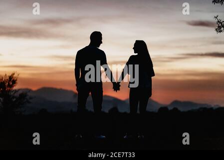 Rückansicht Silhouetten von unkenntlichen Männern und Frauen, die Hände halten und einander anblicken, während sie während des wunderschönen Sonnenuntergangs in den Bergen auf dem Hügel stehen Stockfoto