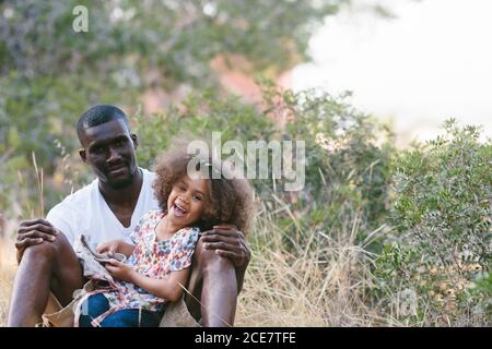 Positive schwarze Mann in legerer Kleidung sitzt mit niedlichen kleinen Tochter auf Runden und lehnte sich auf Baumstamm während der Ausgaben Sonniger Sommertag im Wald Stockfoto