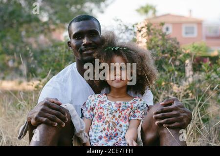 Lächelnder afroamerikanischer Vater sitzt auf der Wiese mit liebenswert froh Tochter zwischen den Beinen und Blick auf die Kamera glücklich in der Landschaft Bereich während des Sommertages Stockfoto