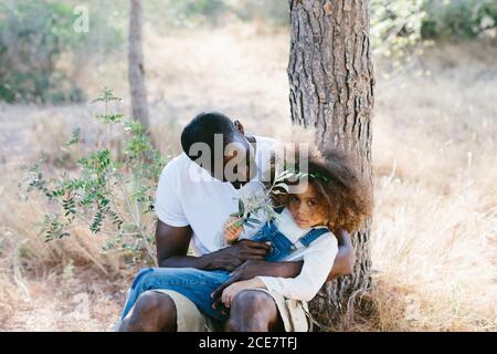 Positive schwarze Mann in legerer Kleidung sitzt mit niedlichen kleinen Tochter auf Runden und lehnte sich auf Baumstamm während der Ausgaben Sonniger Sommertag im Wald Stockfoto