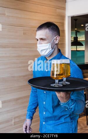 Junger Kellner in Gesichtsmaske und Hemd Trage Tablett mit Gläser frisches leichtes Bier während der Arbeit im Café Stockfoto