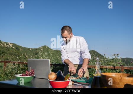 Konzentrierter männlicher Koch im einheitlichen Leserezept auf Laptop und Schneiden von Gemüse beim Kochen Mittagessen auf der Außenterrasse des Restaurants Auf dem Land Stockfoto