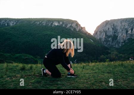 Seitenansicht einer nicht erkennbaren Frau in Sportkleidung, die sich auf das Knie stützt Und Schnürsenkel an Turnschuhen beim Training in den Bergen binden Sonnenuntergang Stockfoto