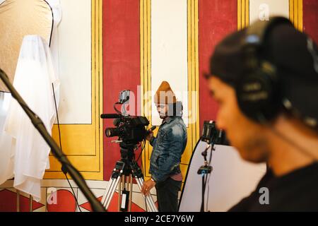 Seriöser Videoanbieter, der Videos auf einer professionellen Kamera auf einem Stativ filmt Nahe Assistent mit Beleuchtung Stockfoto