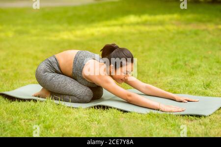 Ruhige asiatische Frau tut Kind Yoga Pose auf Sportmatte Im grünen Park Stockfoto