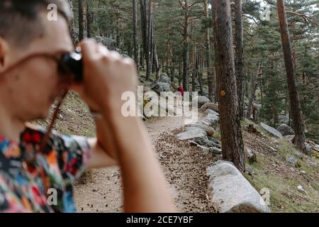 Seitenansicht des männlichen Touristen in buntem Hemd auf dem steht Hang des Berges mit Nadelwald bedeckt und Umwelt beobachten Mit Fernglas beim Wandern in Navacerrada in Spanien Stockfoto