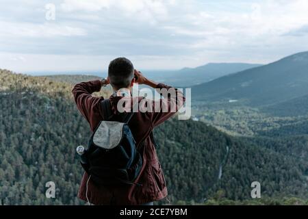 Rückansicht eines nicht erkennbaren männlichen Touristen mit Rucksack auf dem stand Berghang und bewundernde Aussicht auf grüne Hügel während der Beobachtung Landschaft mit Fernglas in Navacerrada in Spanien Stockfoto