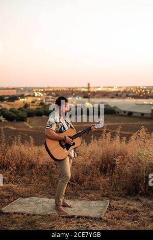Seitenansicht barfuß Hipster Gitarrist in Freizeitkleidung spielen Klassische Gitarre beim Stehen auf dem Teppich in der Nähe von Gras hinter Feld Bei Sonnenuntergang unter ruhigem Himmel Stockfoto