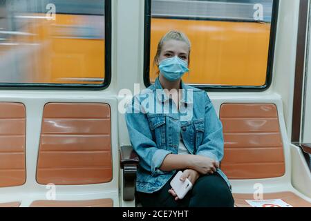 Millennial weibliche Passagier in lässigen Outfit und Schutzmaske suchen In der Kamera allein sitzend in der U-Bahn mit Verbotsschildern Auf Sitzen, um bei einer Coronavirus-Pandemie Abstand zu halten Stockfoto