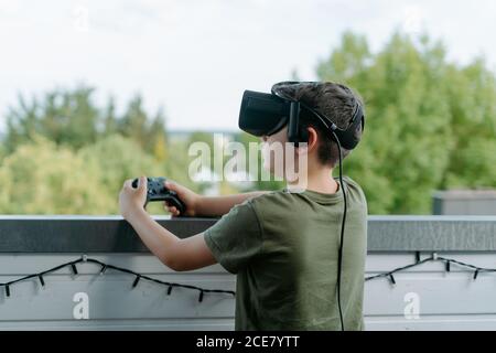 Neugieriger kleiner Junge im T-Shirt, der allein auf dem Balkon steht Und mit VR-Brille und Joystick wegschauen Stockfoto