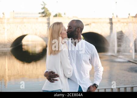 Liebevolle verschiedene Paar in lässigen Outfits umarmen und küssen während Stehen gegen schöne Steinbrücke an sonnigen Tag Stockfoto