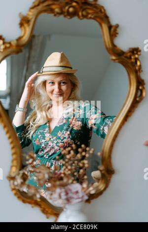 Charmanter Inhalt Frau trägt florale grüne Kleid und trendigen Hut Blick in gemütlichen Spiegel mit goldenem Rahmen, während berühren Hut Stockfoto