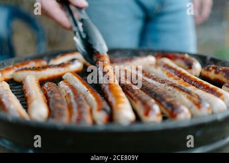 Crop anonyme Person in Jeans Braten köstliche Rinderwürste auf Grillen und Zangen beim Grillen Picknick auf dem Hinterhof Stockfoto