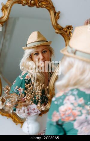Charmanter Inhalt Frau trägt florale grüne Kleid und trendigen Hut Blick in gemütlichen Spiegel mit goldenem Rahmen wegschauen Stockfoto