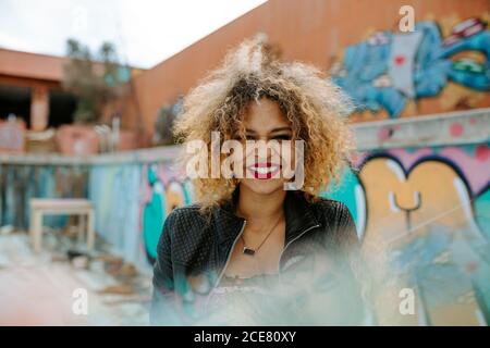Entzückten afroamerikanischen weiblichen Millennial in stilvollem Outfit und mit Afro Frisur steht in der städtischen Umgebung mit Graffiti und suchen Bei der Kamera Stockfoto