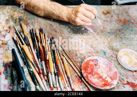 Von oben Ernte anonyme männliche Künstler Hand mit Pinsel oben Schmutzige Arbeitsplatte mit verschiedenen Bürsten und Palette Stockfoto