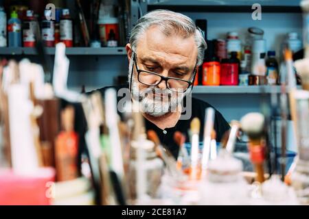 Fokussierte reife Künstler in Brillen arbeiten hinter Sammlung von Pinsel Von verschiedenen Größen und Flaschen mit Farbe in kreativen Studio Stockfoto