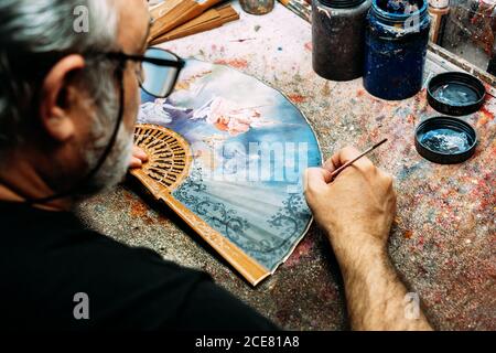 Rückansicht von oben gesichtsloser männlicher Maler mit Pinsel zu Zeichnen Sie auf Handventilator, während Sie am unordentlichen Tisch sitzen und Arbeiten in kreativen Werkstatt Stockfoto