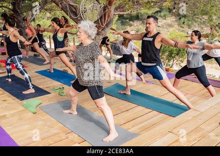 Gruppe von motivierten Tauchern in verschiedenen Altersstufen, die Beine strecken Und Knöchel während der Durchführung Virabhadrasana B Pose während Outdoor-Yoga Klasse Stockfoto