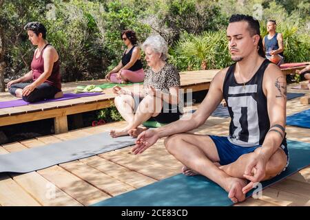 Junger Mann und ältere Frau in einer Gruppe von Menschen meditieren in Lotus-Pose mit geschlossenen Augen während Yoga-Praxis in tropischen Resort an sonnigen Tag Stockfoto